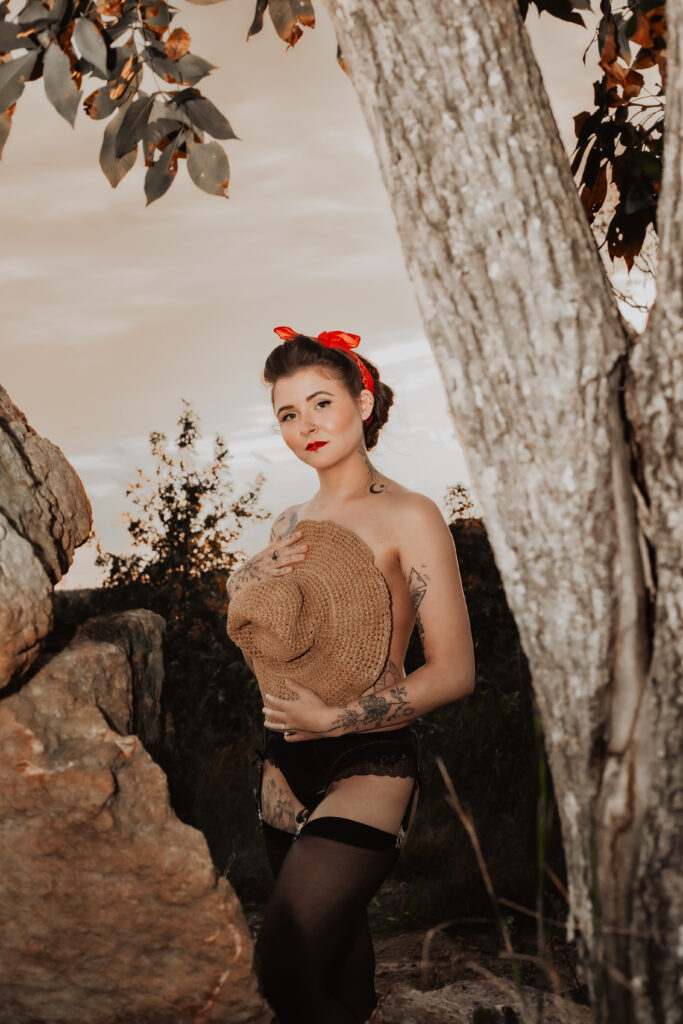 A modern take on a classic pinup girl, shirtless, covered by a straw hat with the sun setting in the background.