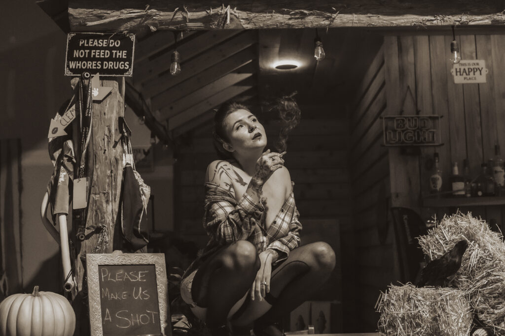 Retro pinup model posing in a rustic bar scene, styled by Lux Verum Productions in Chapin, South Carolina. The sepia-toned image features the model wearing a loose plaid shirt, black stockings, and heels, holding a cigarette with a confident, vintage flair. Surrounded by hay bales, playful signage, and warm wooden textures, the photo captures the timeless charm of classic pinup photography blended with a country-inspired aesthetic.