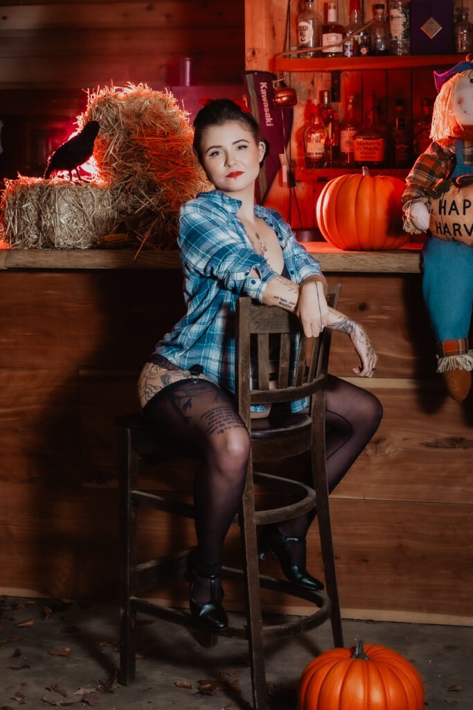 In this captivating image from Lux Verum Productions, a modern pinup model channels vintage allure with a playful edge. Dressed in classic black lingerie, garter belt, and stockings, she poses confidently atop a rustic bar, surrounded by warm wood tones, hay bales, and retro décor. Her red lipstick, victory-roll hairstyle, and subtle smile recall the timeless confidence and flirtatious grace of 1940s pinup art. The soft red lighting adds a sultry yet elegant glow, highlighting how Lux Verum blends nostalgia and sophistication—bringing back the art of tasteful seduction in a way that celebrates femininity, strength, and fun.